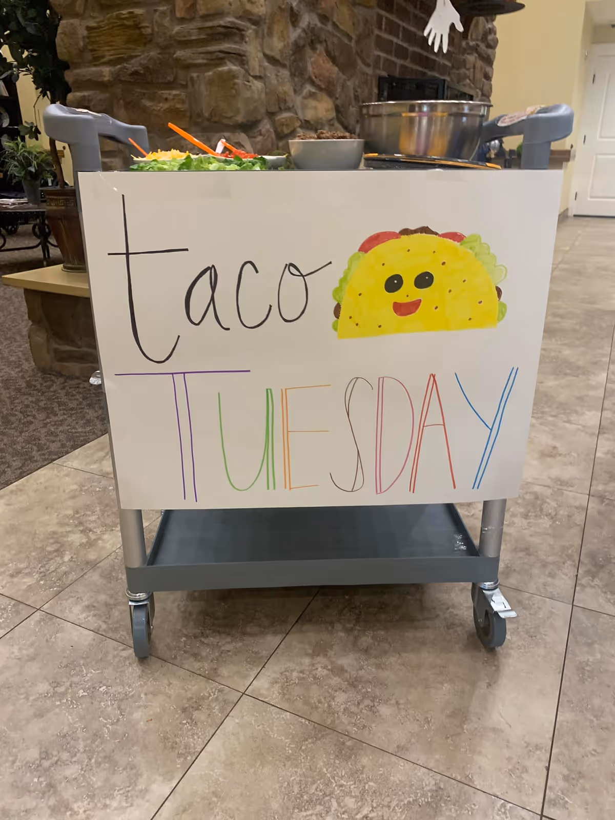 A food cart with a handmade sign that reads 'taco TUESDAY' with a colorful drawing of a smiling taco. The cart has bowls of taco ingredients on top and is positioned on a tiled floor near a stone wall and a hallway.