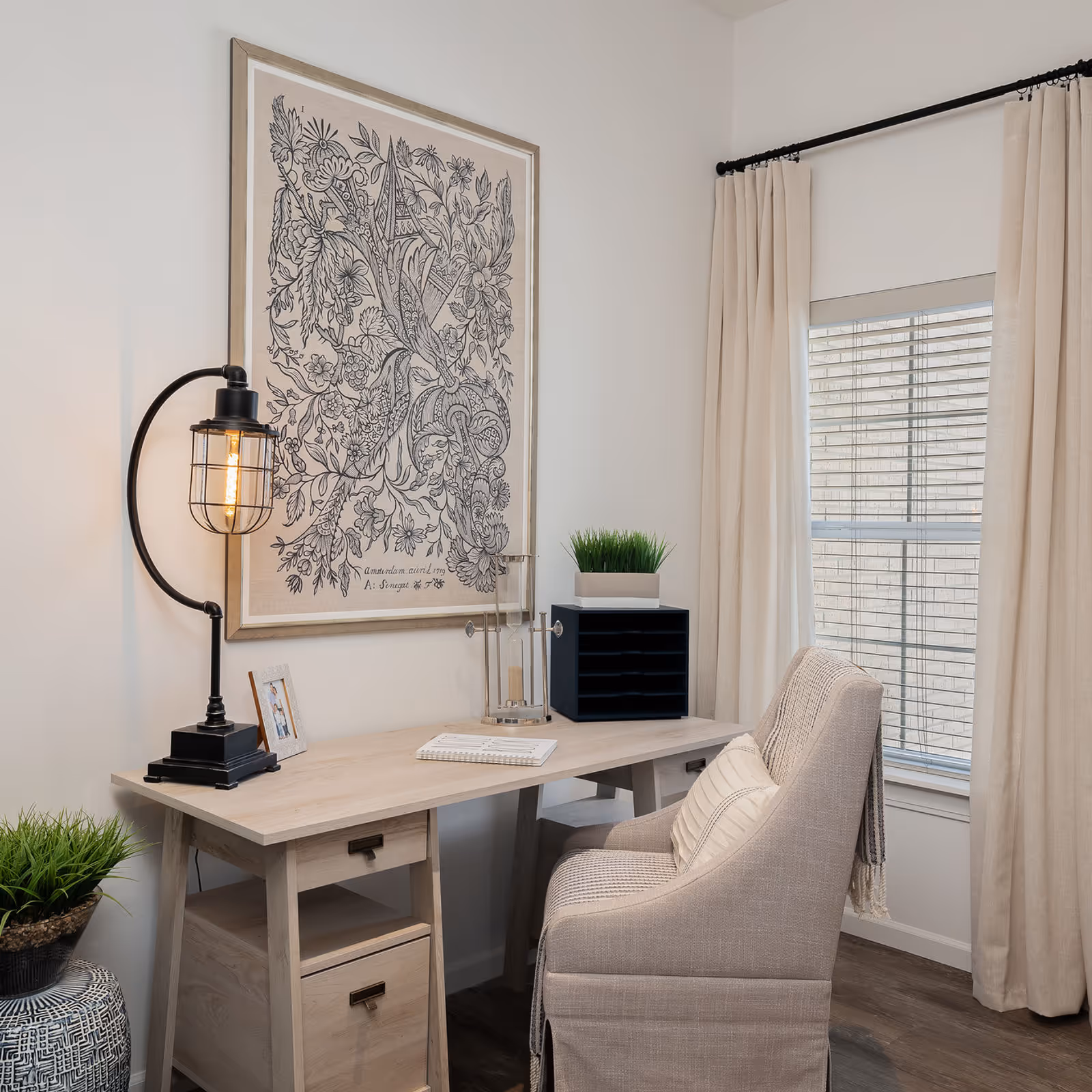 A light wood desk with a lamp, framed botanical artwork, potted plants, and an upholstered chair beside a window with beige curtains.