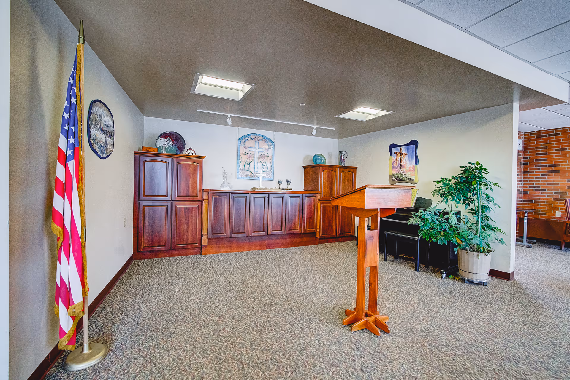 Interior room with a wooden podium in the center, an American flag on a stand to the left, wooden cabinets along the back wall, religious artwork on the walls, a piano with a potted plant beside it, and carpeted flooring.