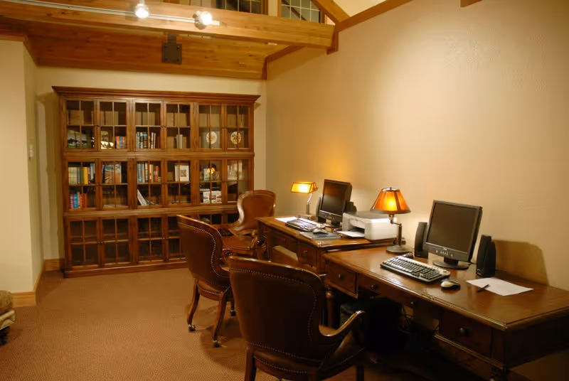 A cozy office or study room with a wooden desk holding two computer monitors, keyboards, a printer, and two table lamps. There are three brown leather chairs in front of the desk. In the background, there is a large wooden bookshelf filled with books and decorative items. The room has a warm, inviting atmosphere with wooden ceiling beams and soft lighting.
