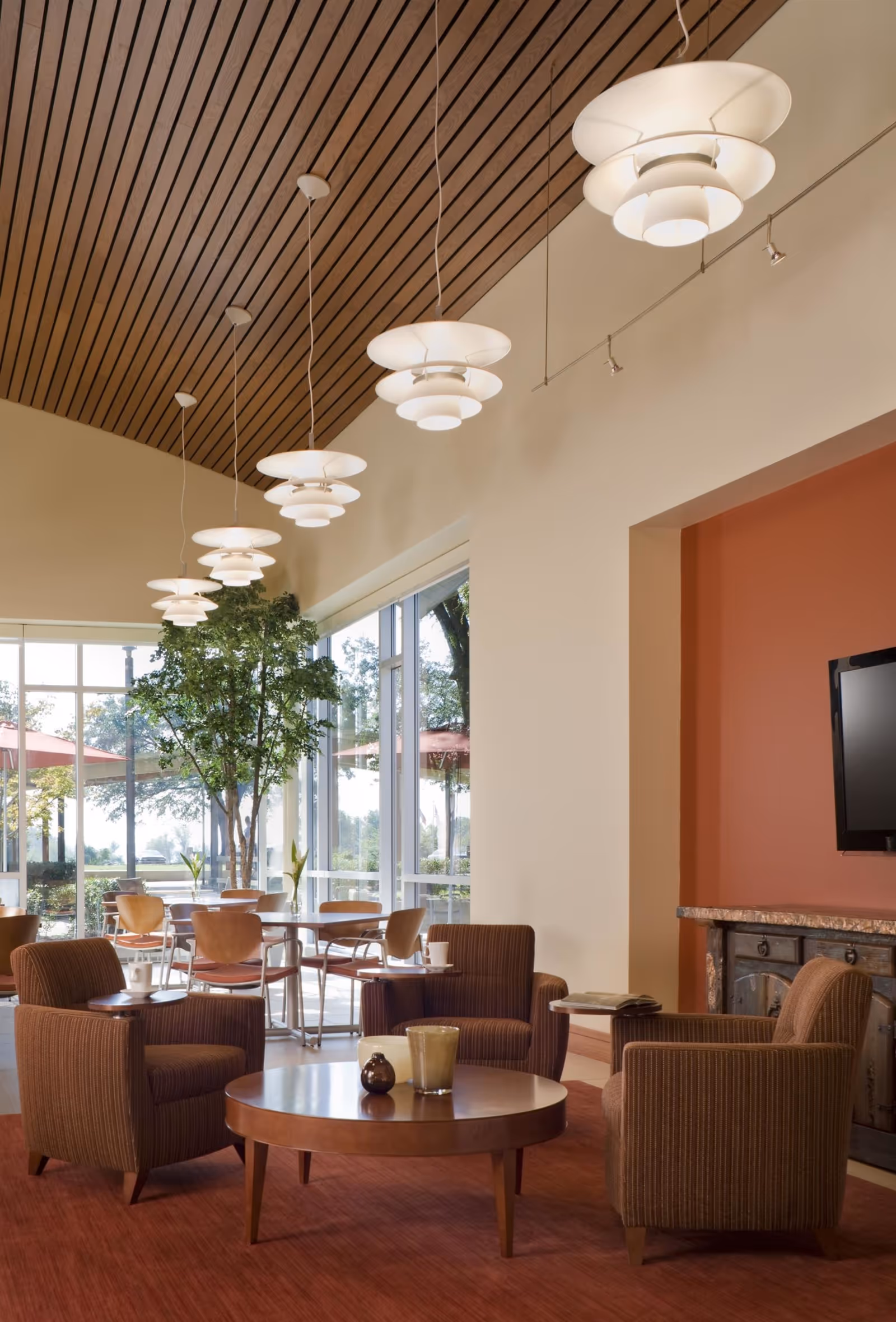 Bright communal lounge with armchairs around a round coffee table, pendant lights overhead, and large windows overlooking outdoor seating.