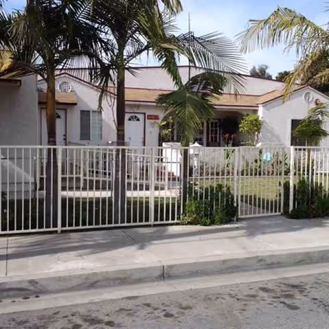 Front exterior of a single-story senior living facility with a white fence and palm trees.