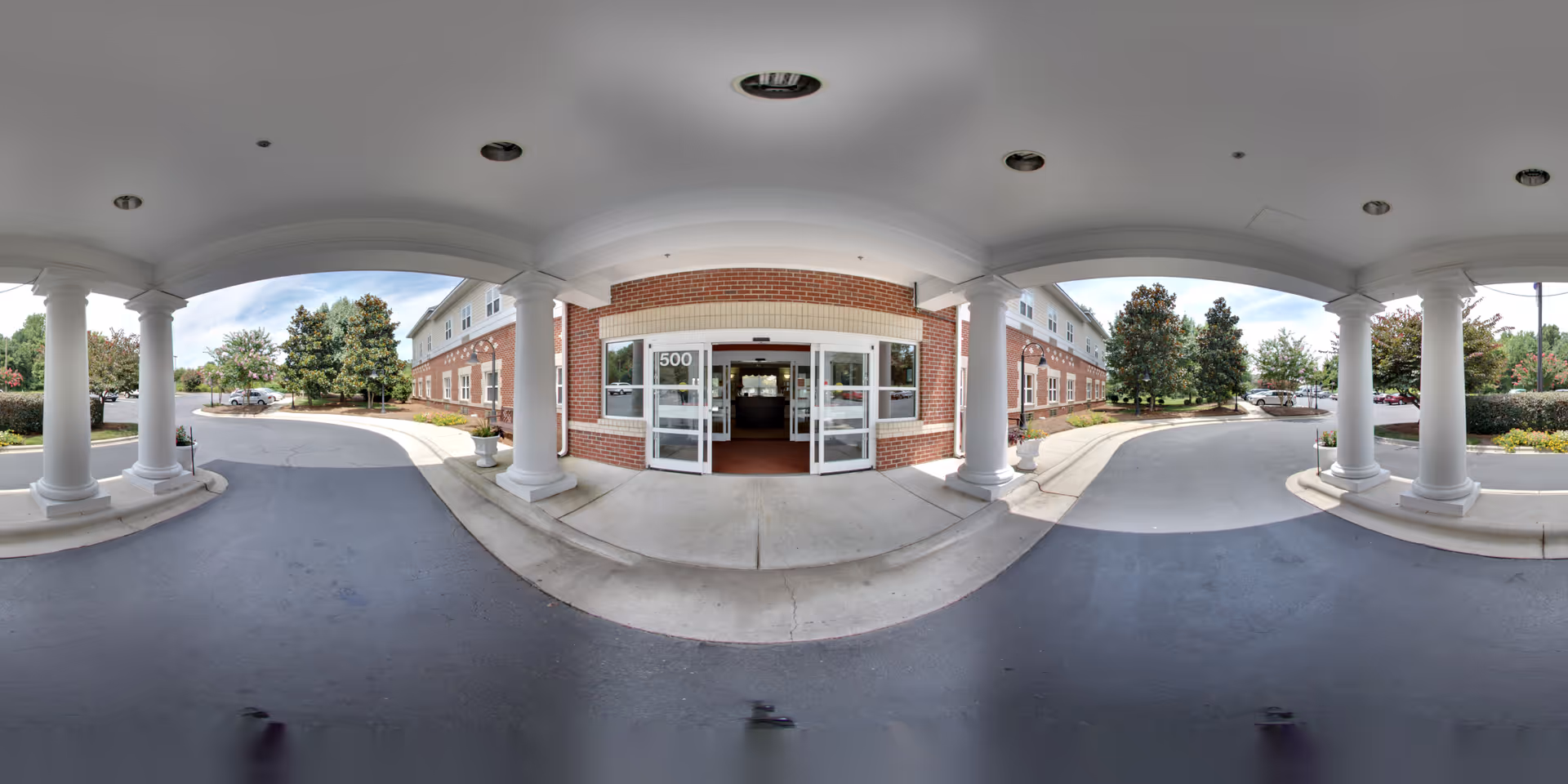 Entrance of a senior living facility with a covered drop-off area supported by white columns. The building has a brick facade with large glass double doors marked with the number 500. Surrounding the entrance are landscaped areas with trees and bushes, and a paved driveway curves around the front.