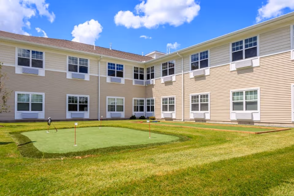 Outdoor courtyard area of a senior living facility with a small putting green and bocce ball court surrounded by a two-story beige building under a blue sky with scattered clouds.