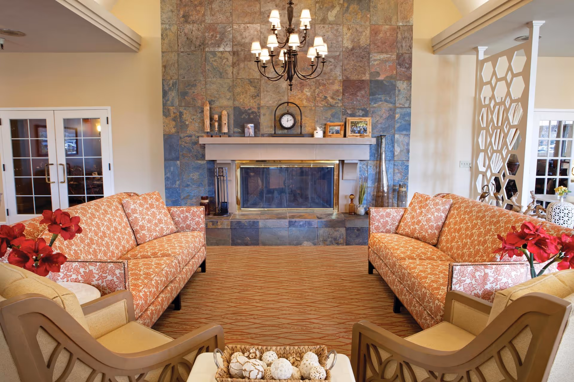 A cozy living room with two patterned orange sofas facing each other, two beige armchairs, a stone fireplace with a clock and decorative items on the mantel, a chandelier hanging from the ceiling, and red flowers on side tables.