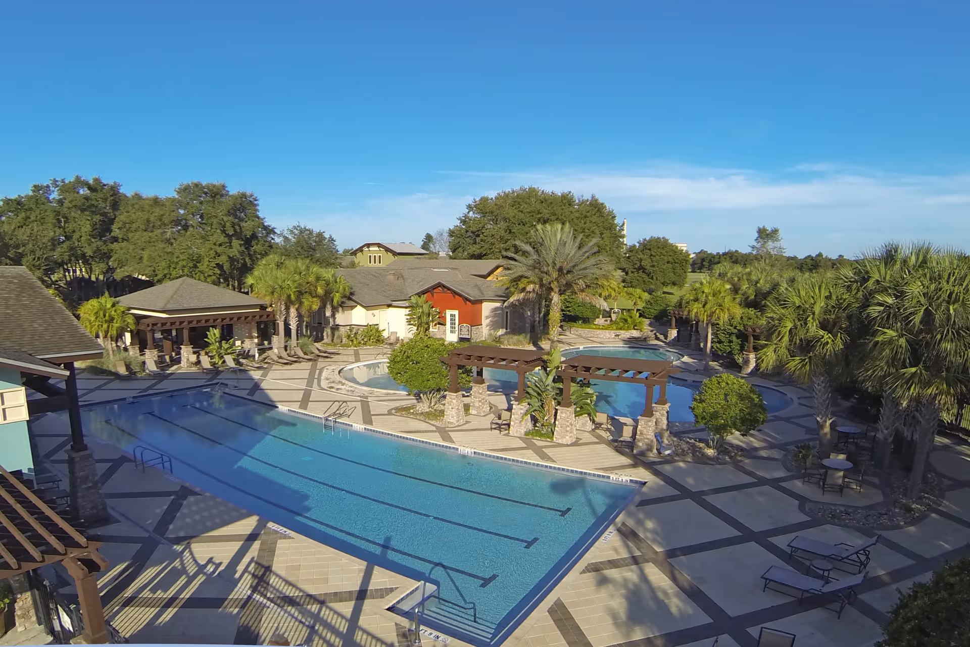 Outdoor view of a senior living facility's pool area with a large swimming pool, a smaller circular pool, palm trees, shaded pergolas, lounge chairs, and tables with chairs under a clear blue sky.