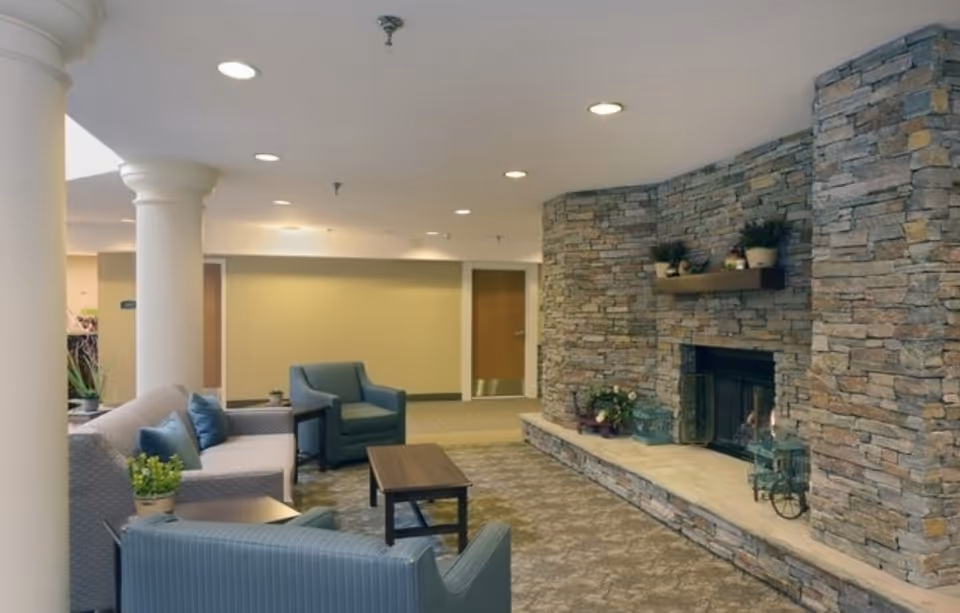 A cozy living room area in a senior living facility with a stone fireplace on the right wall, a wooden mantel with potted plants, a beige couch with blue pillows, two blue armchairs, a wooden coffee table, and large white columns. The room has recessed ceiling lights and a carpeted floor.