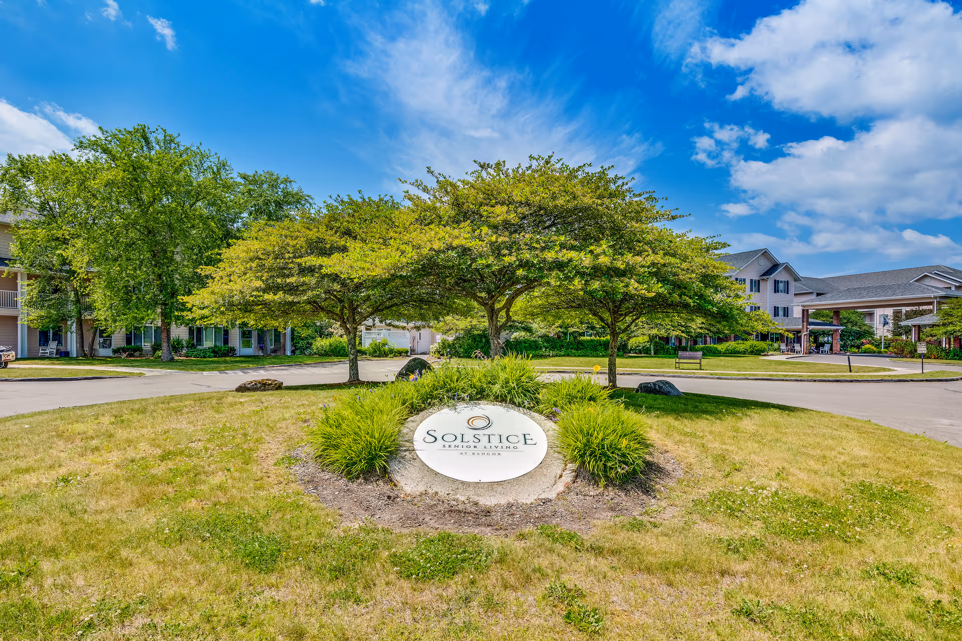 Landscaped circular driveway with a Solstice Senior Living sign and the facility buildings in the background under a blue sky.
