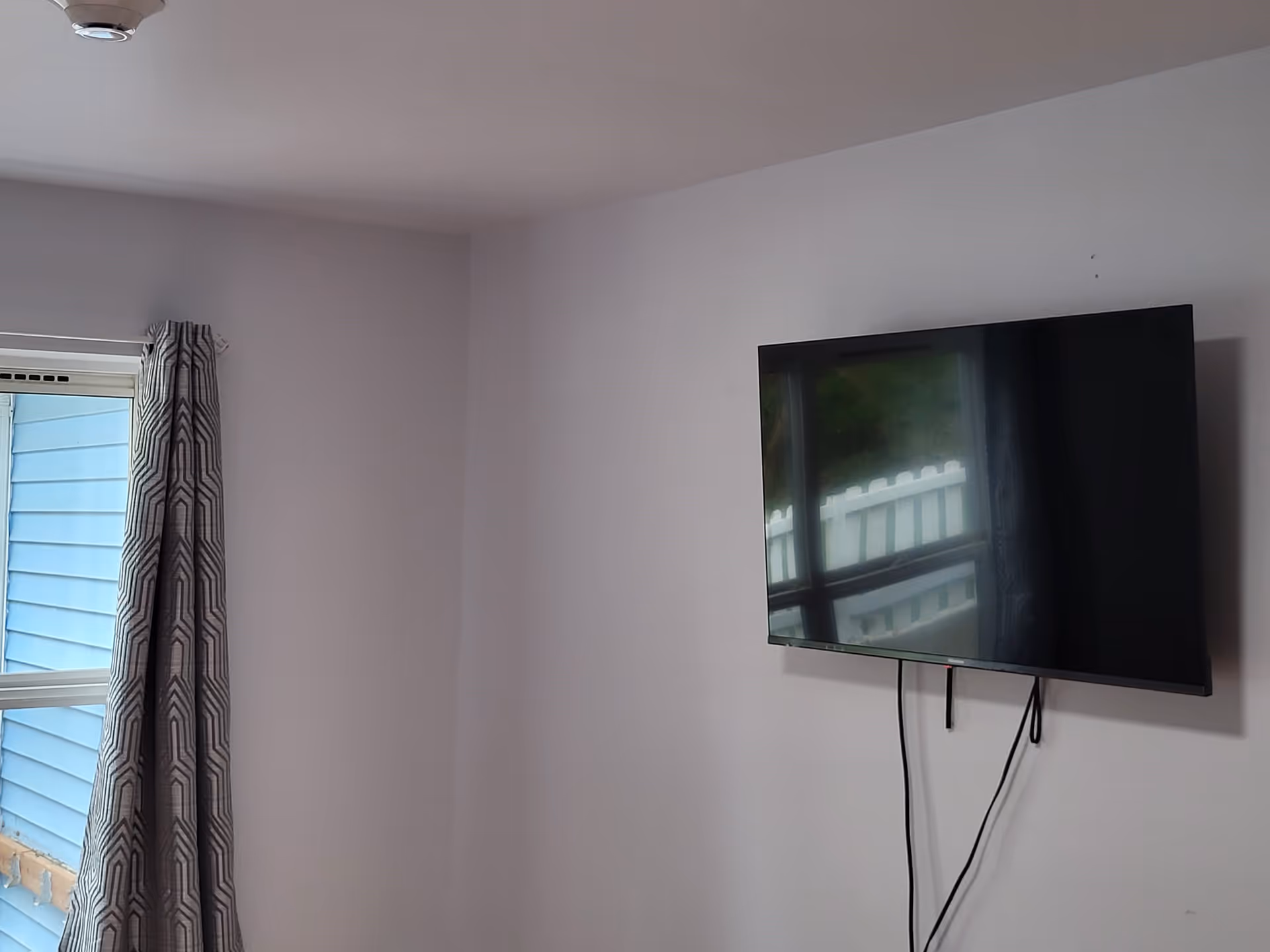 Wall-mounted flat-screen TV on a pale wall next to a window with a patterned curtain.