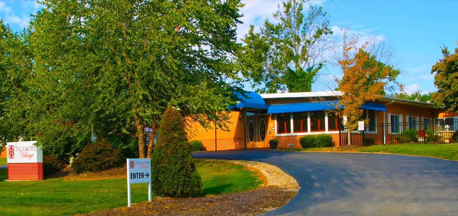 Exterior view of Sycamore Village Assisted Living facility showing a single-story brick building with blue awnings over the entrance and windows. There is a curved driveway leading to the entrance, surrounded by green grass, trees, and shrubs. Two signs are visible on the lawn, one with the facility name and logo, and another directing to the entrance.