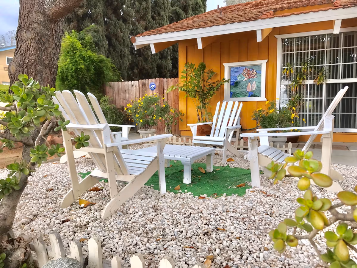 Outdoor seating area with four white wooden chairs arranged around a small white table on a bed of white gravel. The area is surrounded by plants and trees, with a yellow building featuring a window with a floral stained glass decoration in the background.