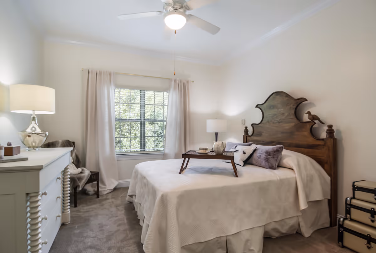 Bright bedroom with a wooden headboard, neatly made bed with a serving tray, dresser, lamps, and a window with curtains.