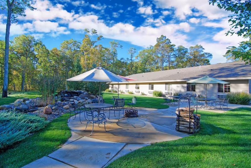 Outdoor patio area at Golden Horizons of Crosslake featuring metal tables and chairs with umbrellas, a fire pit, a stack of firewood, surrounded by green grass, trees, and a building in the background under a partly cloudy blue sky.
