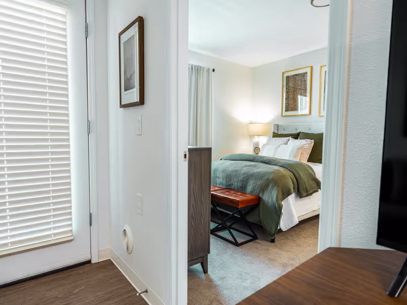 View through a doorway into a bedroom with a neatly made bed featuring green bedding and multiple pillows. A brown bench is positioned at the foot of the bed. The room has light-colored walls, a lamp on a bedside table, framed artwork above the bed, and a window with light curtains. In the foreground, there is a door with white blinds and part of a wooden surface, possibly a table or dresser.