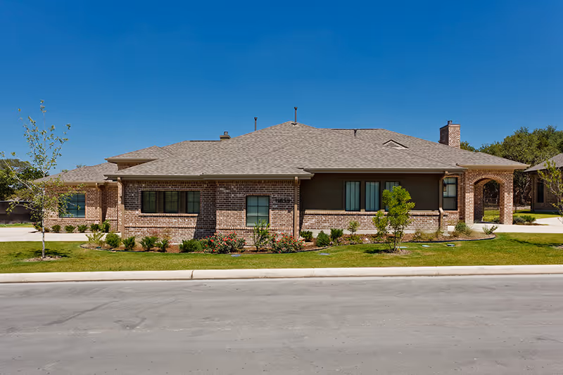 Single-story brick building with a gray shingled roof, several windows, and a small landscaped area with bushes and young trees in front, under a clear blue sky.