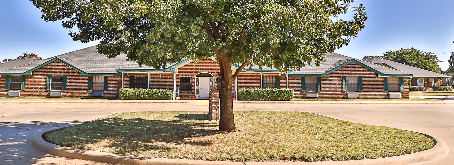 Front exterior view of a single-story brick building with green trim and multiple windows, surrounded by a paved driveway and a grassy area with a tree in the center.