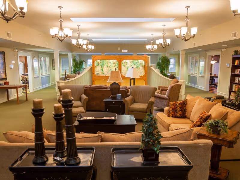 A spacious and well-lit living room area in Buchanan Meadows featuring multiple comfortable sofas and armchairs arranged around coffee tables. The room is decorated with lamps, plants, and candles, with chandeliers hanging from the ceiling. There are large windows and a bookshelf visible in the background.