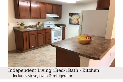 A compact kitchen with dark wood cabinets, a white stove and refrigerator, and a countertop bar holding a bowl of fruit.