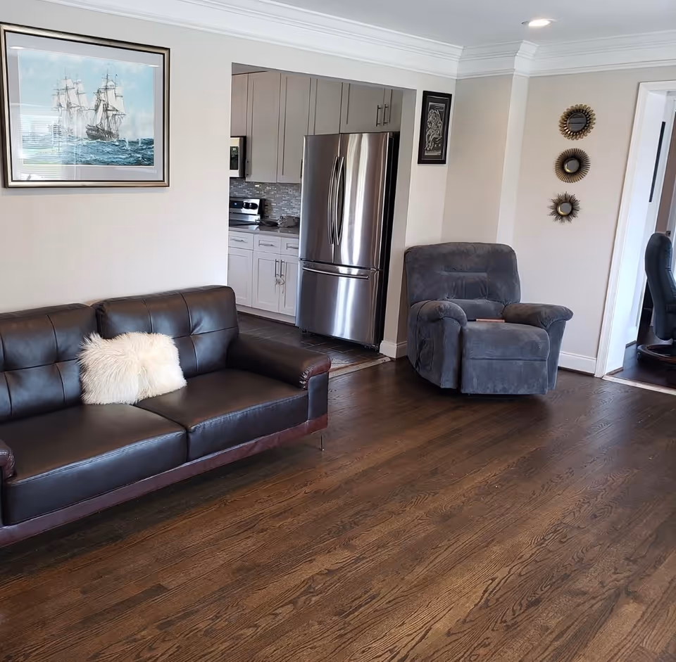 Interior view of a living room with a dark brown leather sofa with a white fluffy pillow, a gray recliner chair, and wooden flooring. The room opens into a kitchen area with white cabinets and a stainless steel refrigerator. Wall decorations include a framed painting of a sailing ship and three round decorative mirrors.