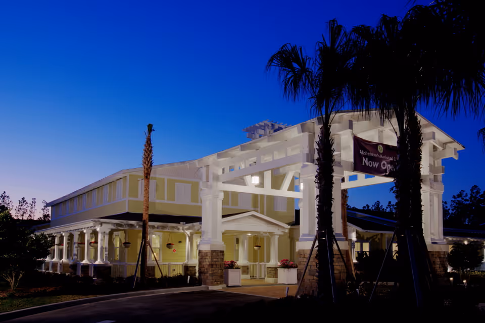 Exterior view of a senior living facility building at dusk with a large white entrance structure supported by columns and palm trees. The building is light yellow with white trim and has a sign that reads 'Alzheimer's Assisted Living Now Open'.