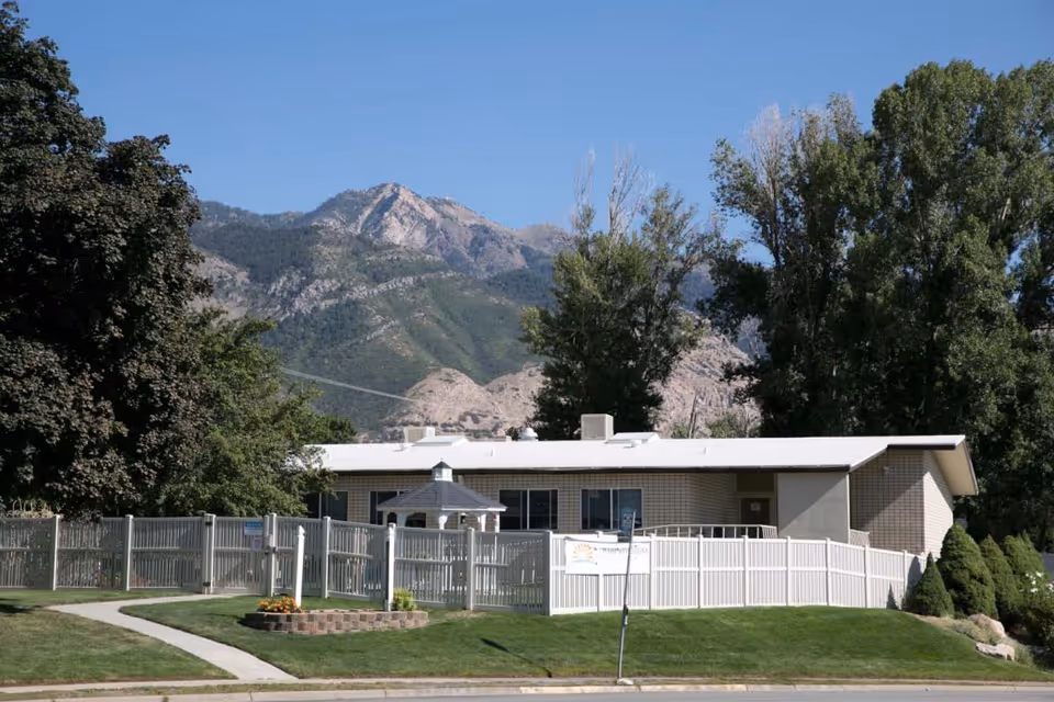 Single-story building with a white roof and beige brick walls surrounded by a white fence. There is a small gazebo inside the fenced area and well-maintained green grass with a flower bed near the sidewalk. Large trees and a mountainous landscape are visible in the background under a clear blue sky.