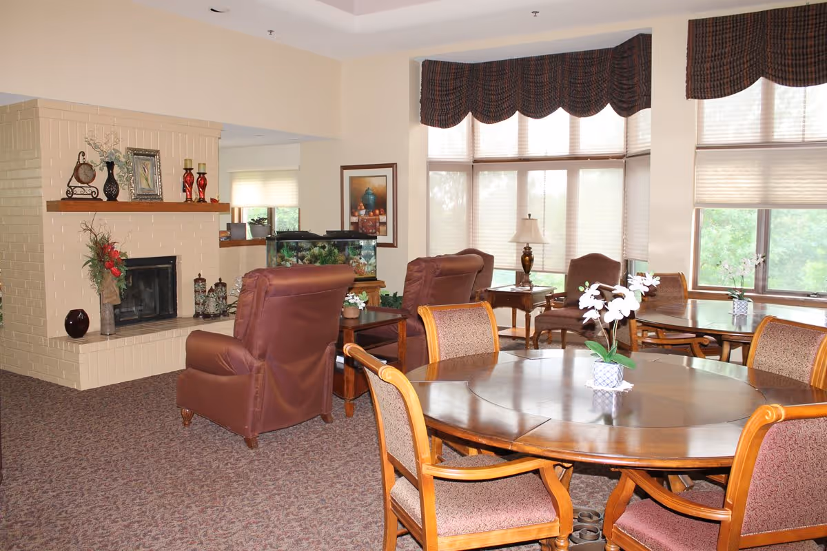 A cozy living room area with a beige brick fireplace decorated with vases and candles. There are two brown leather armchairs facing the fireplace and a wooden round table with four upholstered chairs in the foreground. Large windows with valances and blinds let in natural light, and there is a fish tank and framed artwork on the walls.