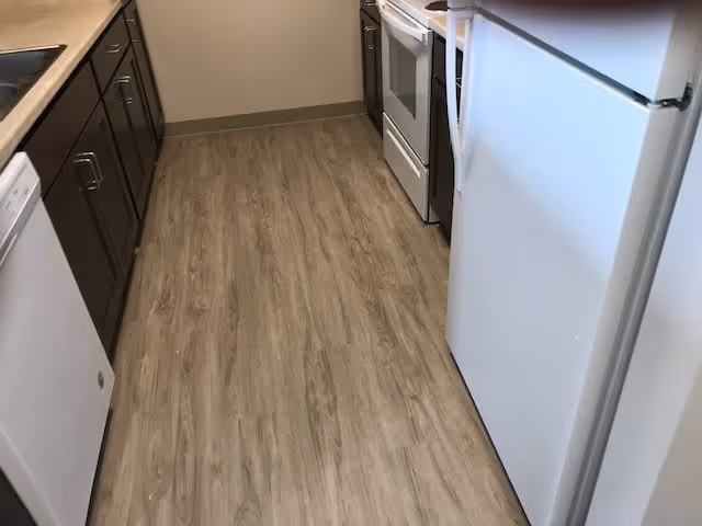 Narrow kitchen with wood-patterned flooring, dark brown cabinets on the left, a white dishwasher, a white refrigerator on the right, and a white stove and oven at the far end.