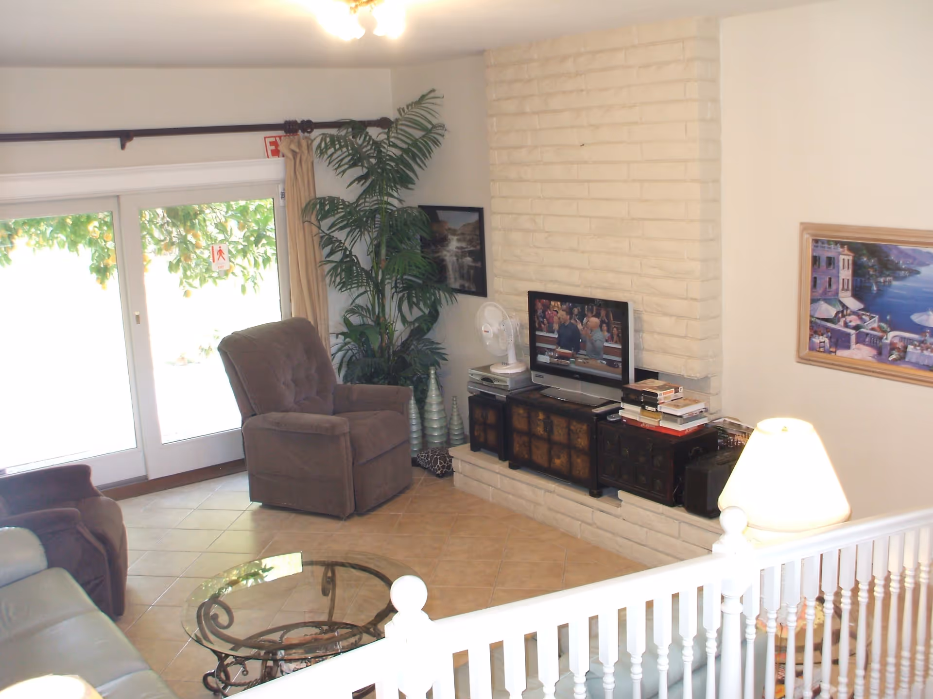 A cozy living room with a beige tiled floor, a brown armchair, a glass coffee table with a decorative metal base, and a TV on a wooden cabinet set against a white brick fireplace. There is a large green plant in the corner near sliding glass doors with beige curtains, a table lamp on the right side, and framed artwork on the walls.