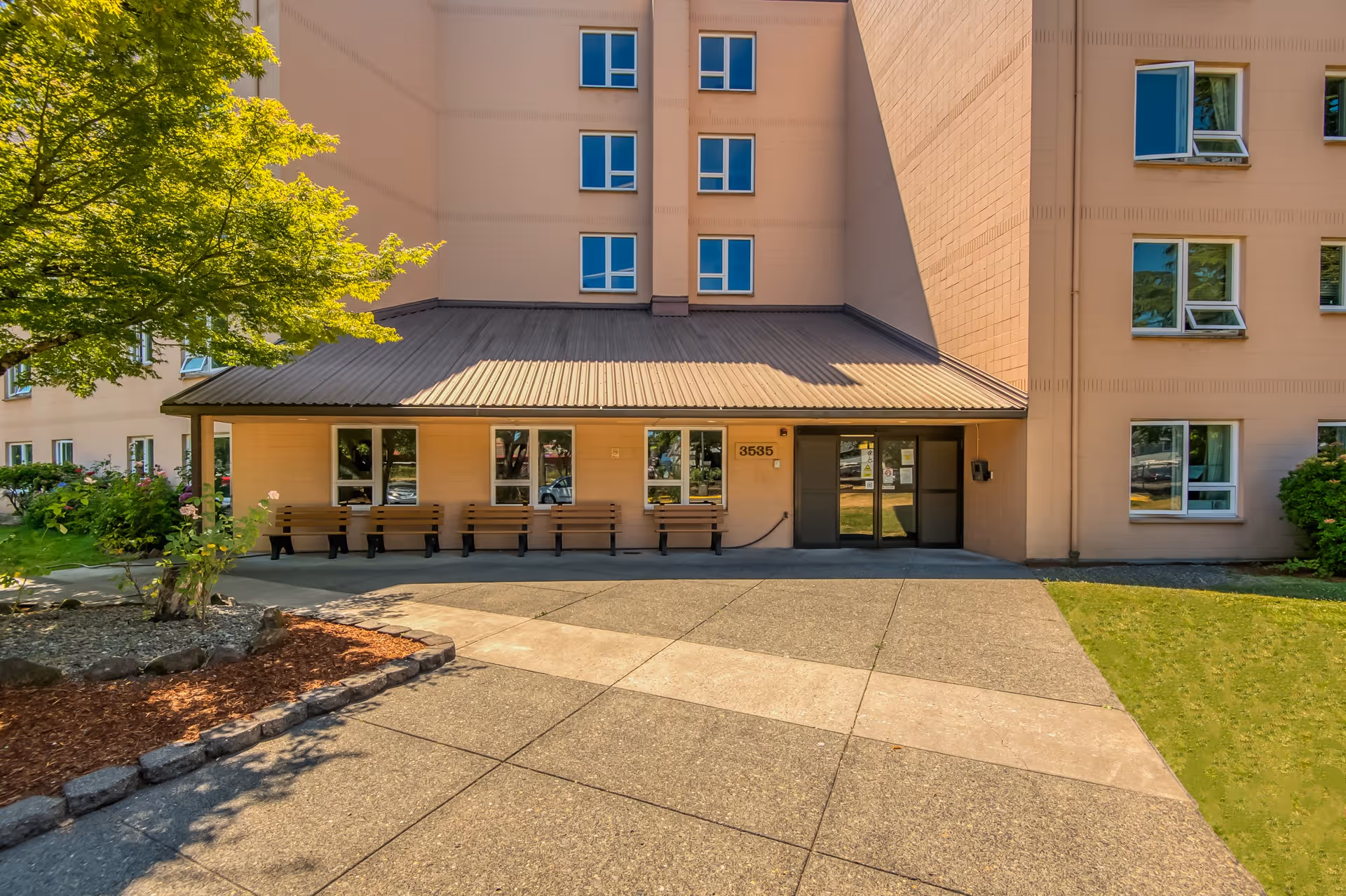 Exterior view of the entrance to a multi-story building with a covered porch area featuring several wooden benches. The building is light peach colored with multiple windows, and there is a tree and landscaped area with mulch and rocks to the left. The entrance has double glass doors and the number 3535 is displayed near the door.