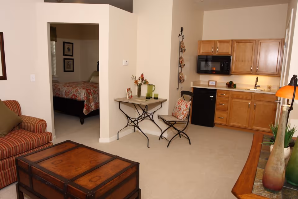 Interior view of a senior living facility apartment showing a small kitchen area with wooden cabinets, a microwave, a mini fridge, and a sink. Adjacent to the kitchen is a small table with two chairs, one with a decorative pillow. A striped armchair and a wooden chest-style coffee table are visible in the foreground. An open doorway leads to a bedroom with a bed covered in a floral bedspread and framed pictures on the wall.