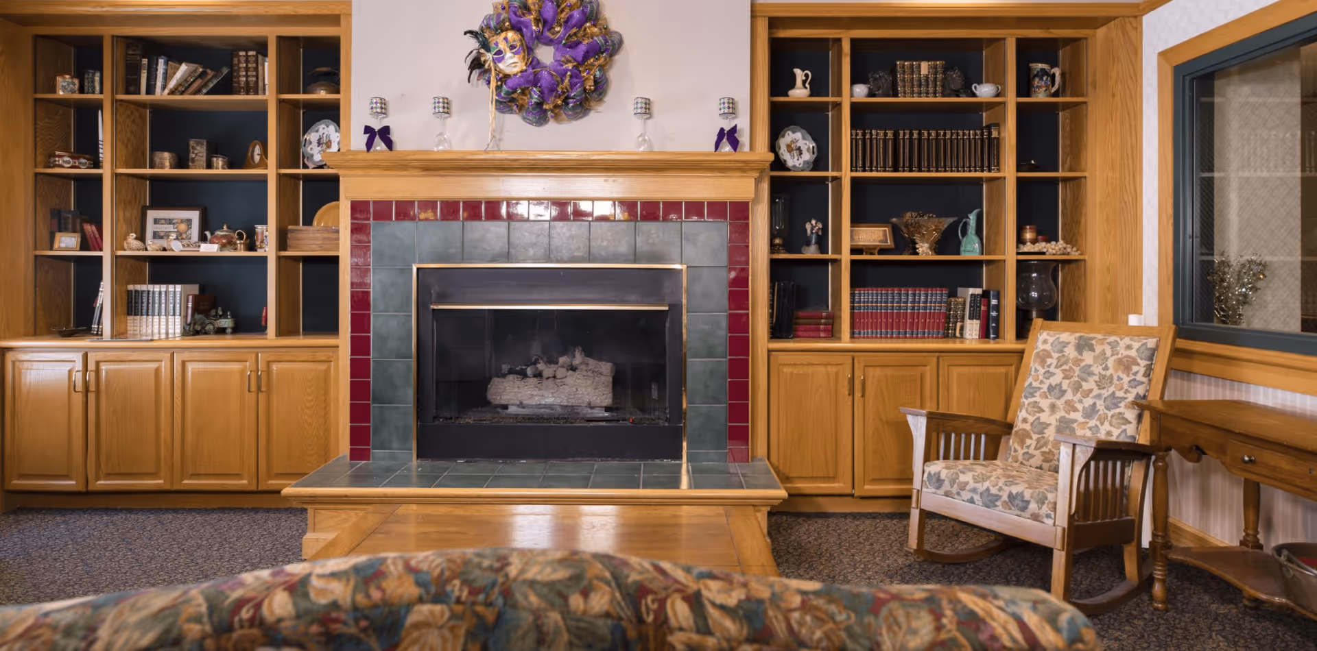 Cozy living room with a fireplace surrounded by green and red tiles, flanked by wooden built-in bookshelves filled with books and decorative items. A floral patterned armchair and a wooden side table are placed near a window with a mesh screen.