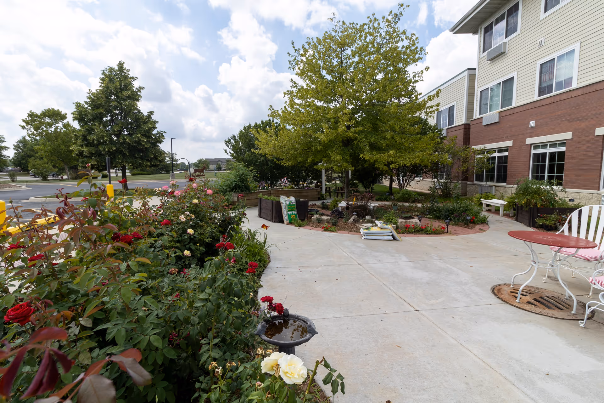 Outdoor patio area at StoryPoint Romeoville featuring a concrete walkway surrounded by flower beds with blooming red and white roses, green trees, and shrubs. There is a small round table with white metal chairs on the right side near the building. The building exterior has beige siding and red brick with several windows. The sky is partly cloudy.