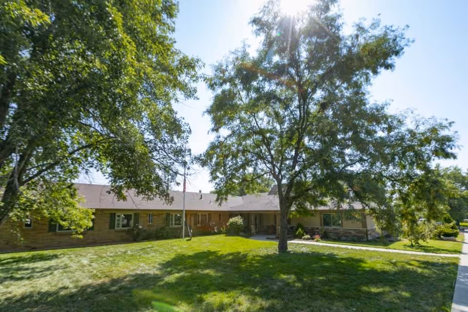 Front exterior of a single-story brick care facility with a grassy lawn, mature trees, a flagpole, and a sidewalk.