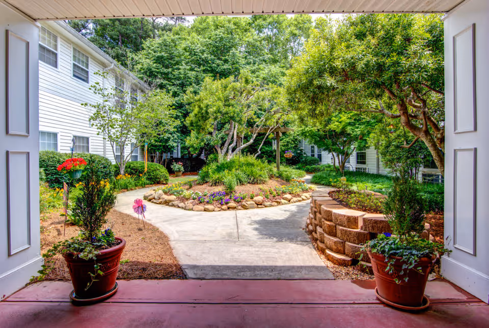 View from a covered walkway looking out onto a landscaped garden courtyard with a circular flower bed surrounded by rocks, various trees, shrubs, and potted plants on either side of the walkway. White residential buildings with multiple windows are visible on both sides.