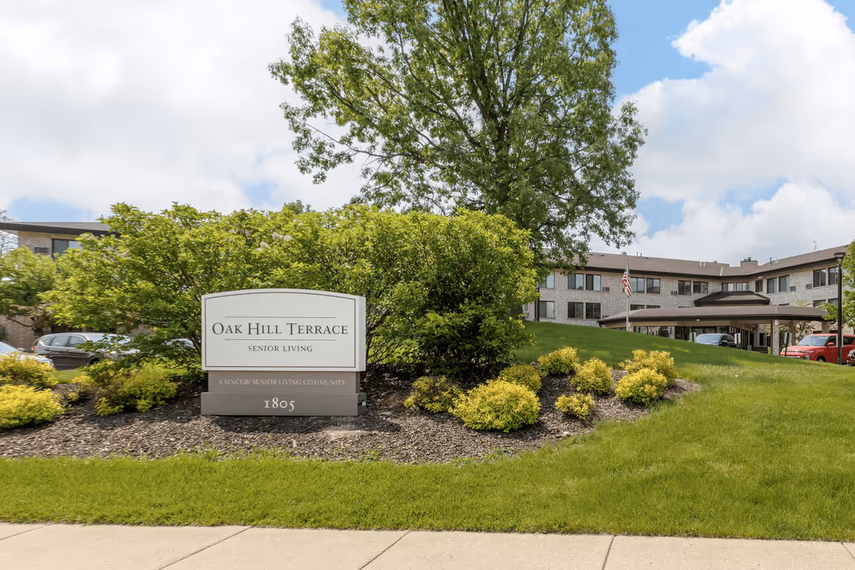 Exterior view of Oak Hill Terrace Senior Living facility with a sign in front surrounded by green bushes and grass, and a multi-story building in the background under a partly cloudy sky.