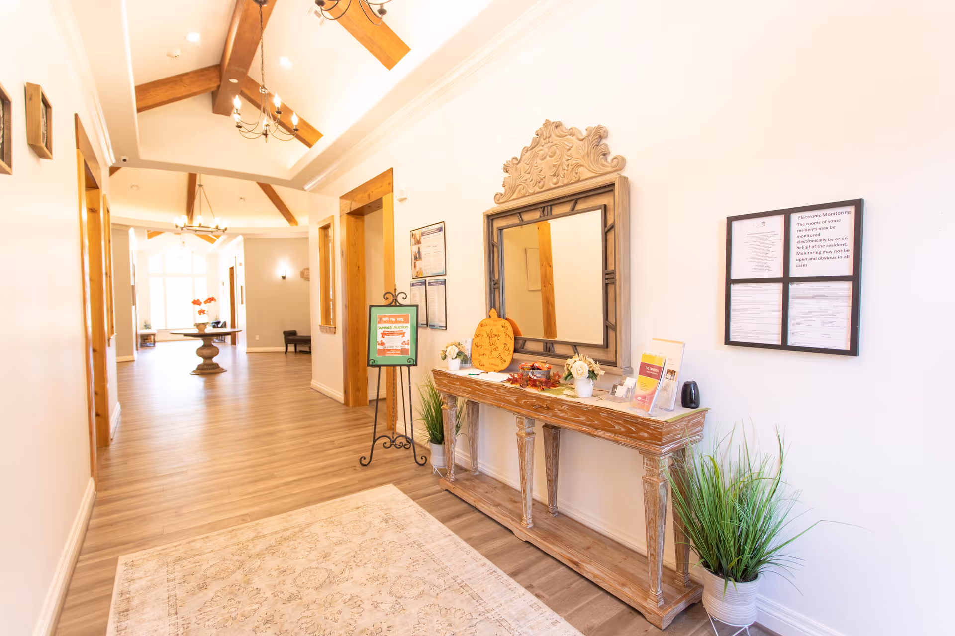 Bright, wide interior hallway with a wooden console table and large mirror, decorative plants, a rug, and exposed ceiling beams.