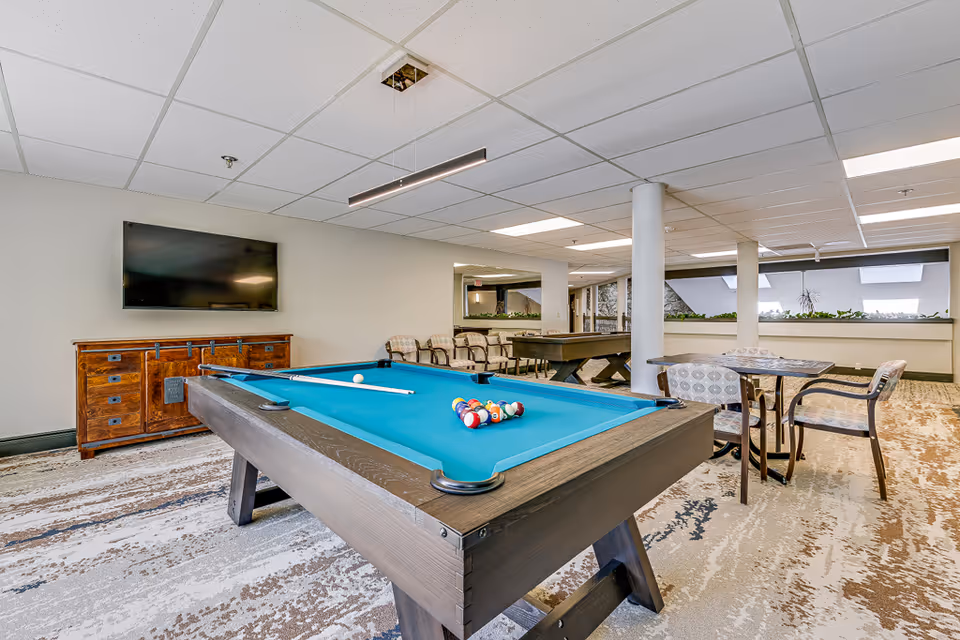A spacious recreational room featuring a pool table with billiard balls and cue sticks, a mounted flat-screen TV on the wall above a wooden cabinet, a small table with four patterned chairs, and another game table in the background. The room has a patterned carpet, white walls, and a drop ceiling with fluorescent lighting.