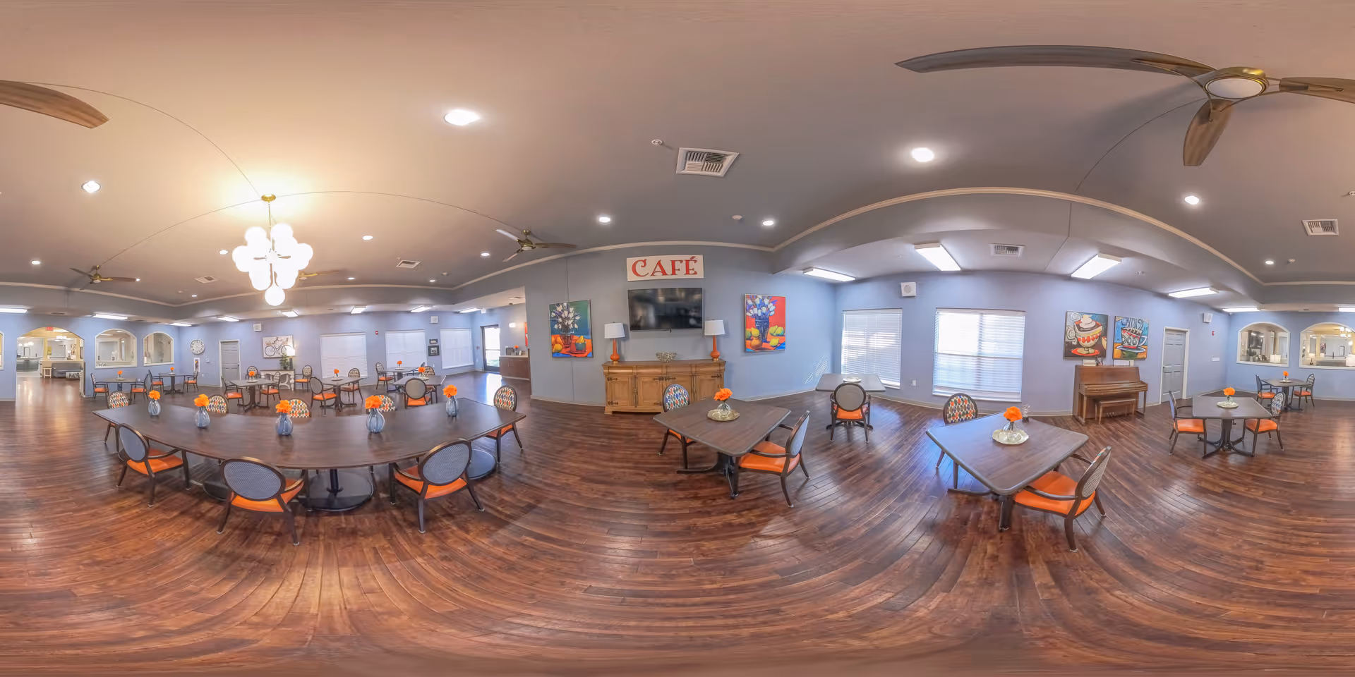 Spacious dining/cafe room with multiple tables and chairs with orange cushions, wood floors, ceiling fans, chandeliers, and a 'CAFÉ' sign on the far wall.