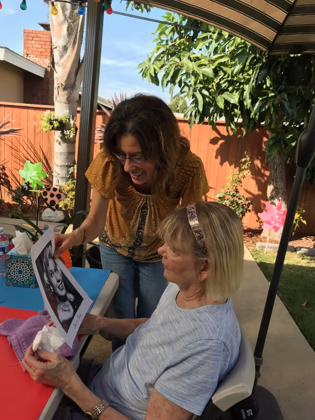 An elderly woman sitting outdoors at a table under a canopy, holding a black and white photo while a younger woman stands beside her smiling and looking at the photo. The setting includes a wooden fence, green plants, and colorful pinwheels in the background.