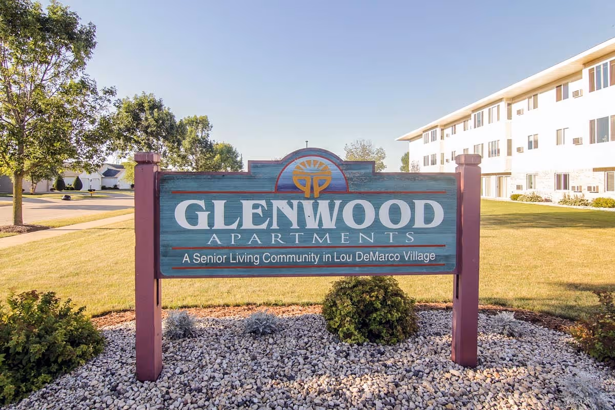 Outdoor view of a wooden sign for Glenwood Apartments, a senior living community in Lou DeMarco Village, with a multi-story white building and green lawn in the background under a clear blue sky.
