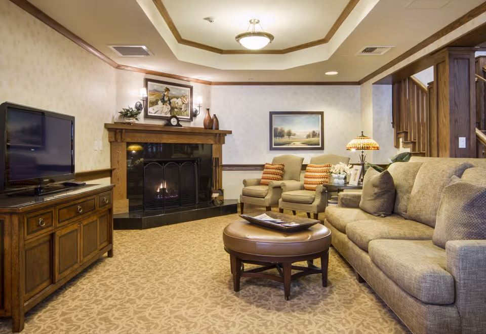 A cozy living room with a beige sofa, two armchairs with orange striped pillows, a round leather ottoman, a wooden TV stand with a flat-screen TV, and a fireplace with a wooden mantle. The room has patterned carpet, framed artwork on the walls, and a decorative lamp on a side table near the armchairs.