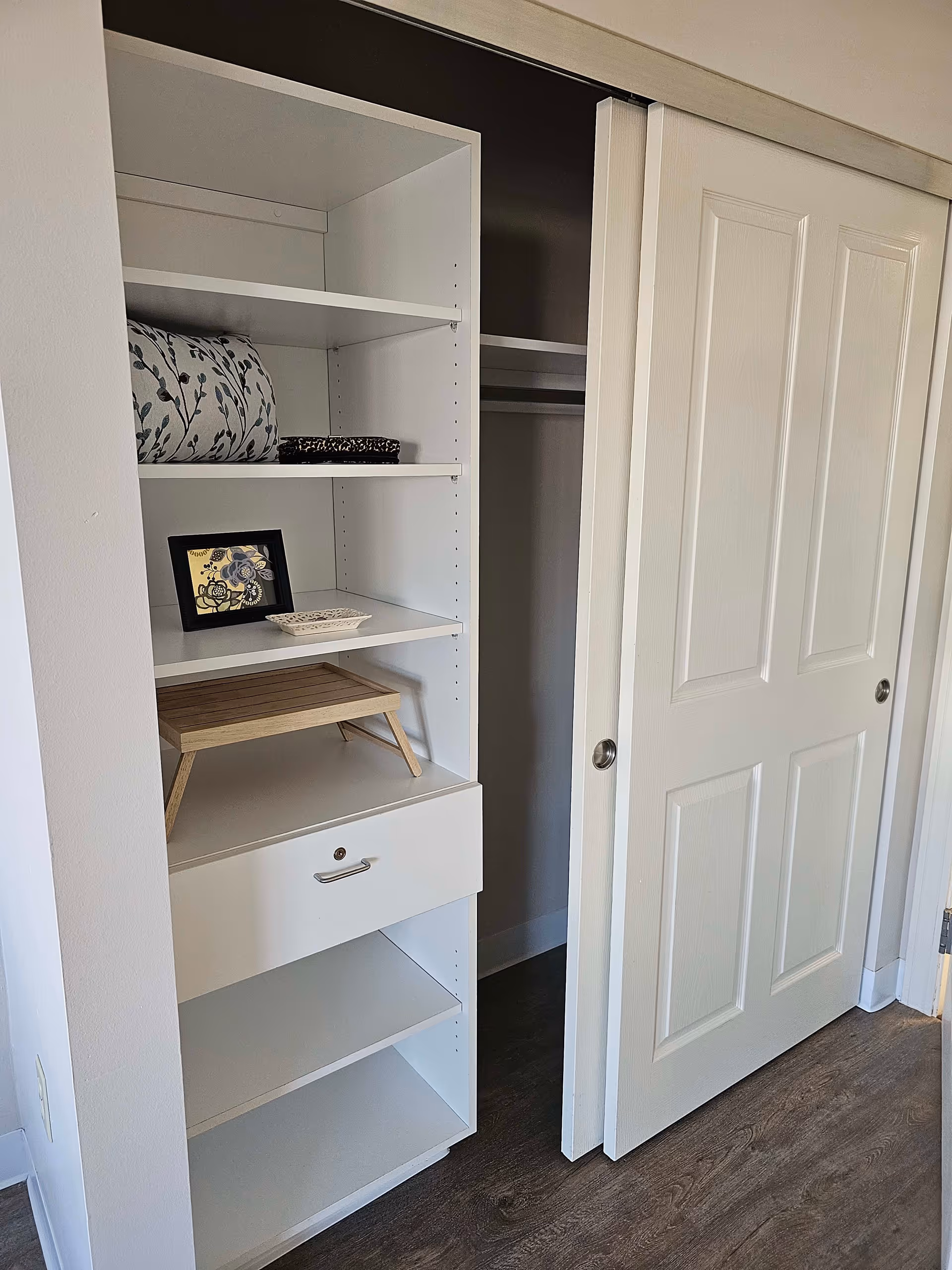 A white built-in shelving unit with several shelves and a drawer, containing a folded blanket, a small framed picture, a decorative dish, and a wooden tray. Next to the shelving unit is a closet with white sliding doors, one door partially open, revealing a hanging rod inside. The floor is wood, and the walls are painted white.