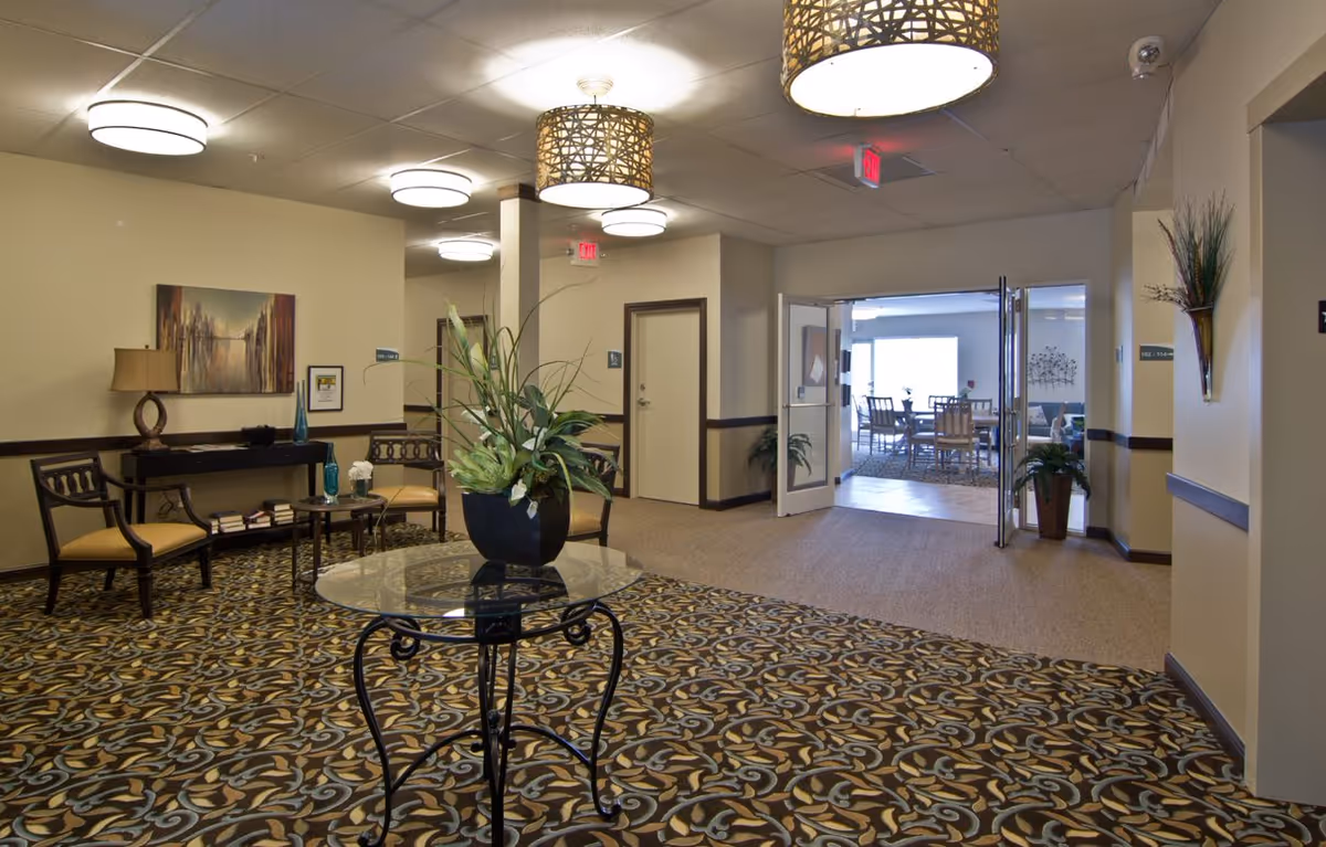 Interior view of a senior living facility lobby area with patterned carpet, a round glass table with a plant centerpiece, chairs along the wall, a console table with a lamp and decorative items, and double doors leading to a dining area with tables and chairs.