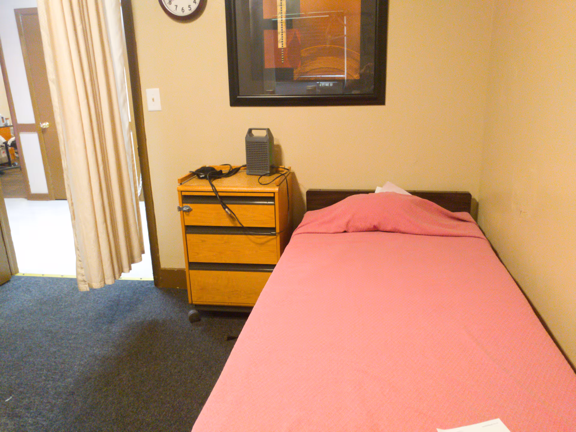 Small single-bed room with a pink bedspread, wooden bedside cabinet, clock and framed artwork on the wall.