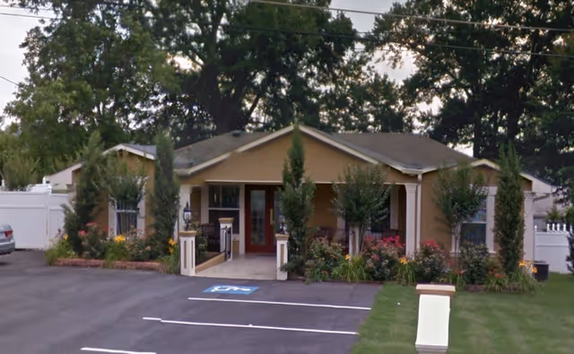 Single-story building with a beige exterior and a covered entrance, surrounded by small trees and flowering plants. There is a paved parking area in front with a handicapped parking space marked. The building is set against a backdrop of tall trees and a white fence on the sides.