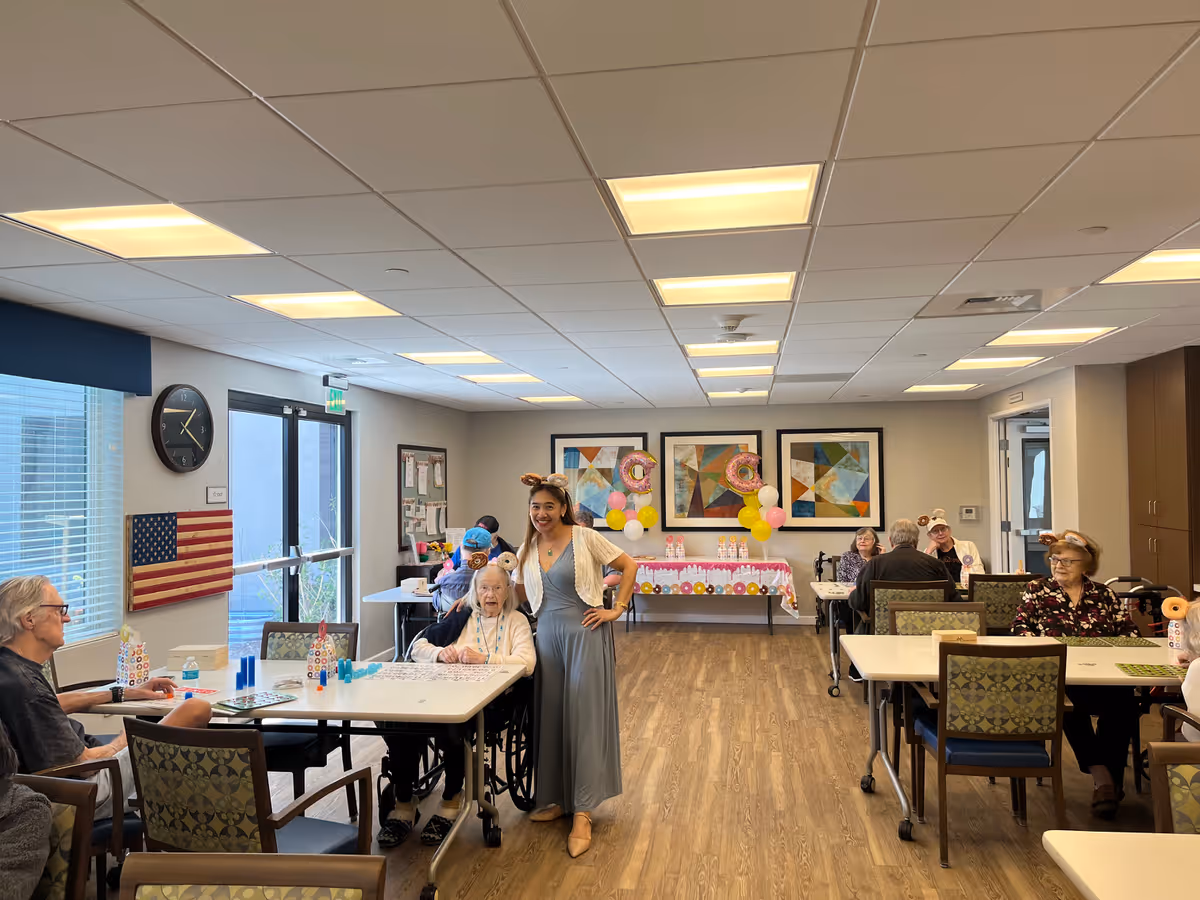 A group of elderly people sitting at tables in a well-lit activity room with wood flooring. A younger woman wearing a long dress and animal ear headband stands smiling next to an elderly woman in a wheelchair. The room has colorful abstract paintings on the wall, balloons, and a table with refreshments in the background. An American flag and a clock are visible on the left wall.