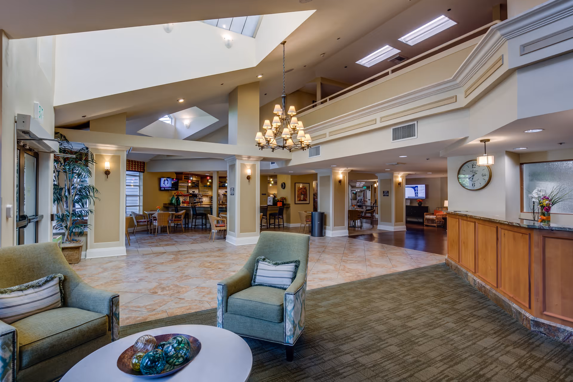 Spacious and well-lit senior living facility common area with comfortable seating including two green armchairs around a round table with decorative glass spheres. The area features a chandelier hanging from a high ceiling with skylights, a reception desk with a clock on the wall, and a dining area with tables and chairs in the background. The decor includes plants and warm lighting, creating a welcoming atmosphere.