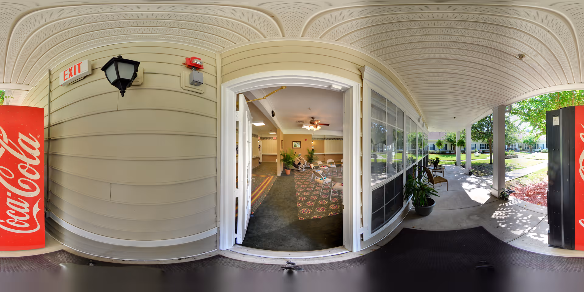 Covered outdoor walkway leading to an open doorway into a common area with chairs and plants inside. There are Coca-Cola vending machines on both sides of the walkway, and the area outside has trees and a garden with a pathway. The ceiling of the walkway is white with a curved design, and there is an exit sign above the door.