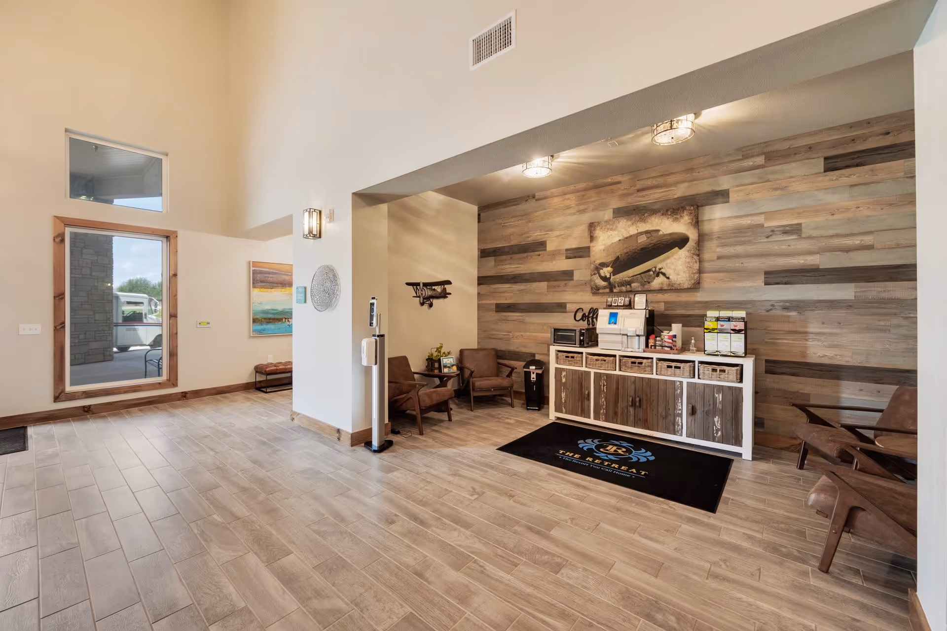 A spacious interior area with light wood flooring and a high ceiling. There is a seating area with brown chairs and a small table. A wooden cabinet against a wood-paneled wall holds a coffee machine, microwave, and various coffee supplies. A black mat in front of the cabinet reads 'The Retreat'. A large window and a glass door are visible on the left side, letting in natural light.