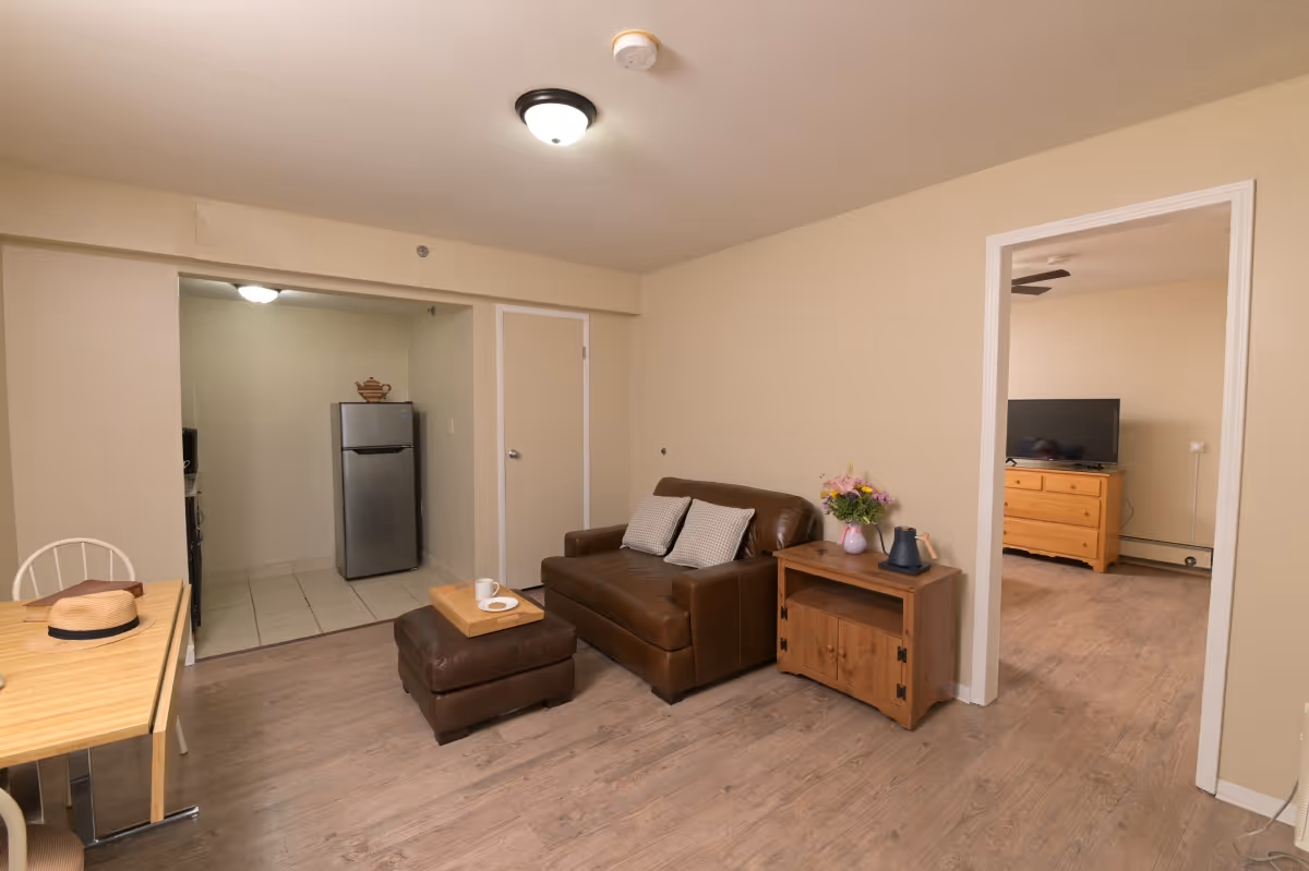Small living area with a brown leather loveseat and ottoman, wooden side table with flowers, dining table, and a view into a kitchenette with a stainless refrigerator and an adjacent bedroom with a TV.